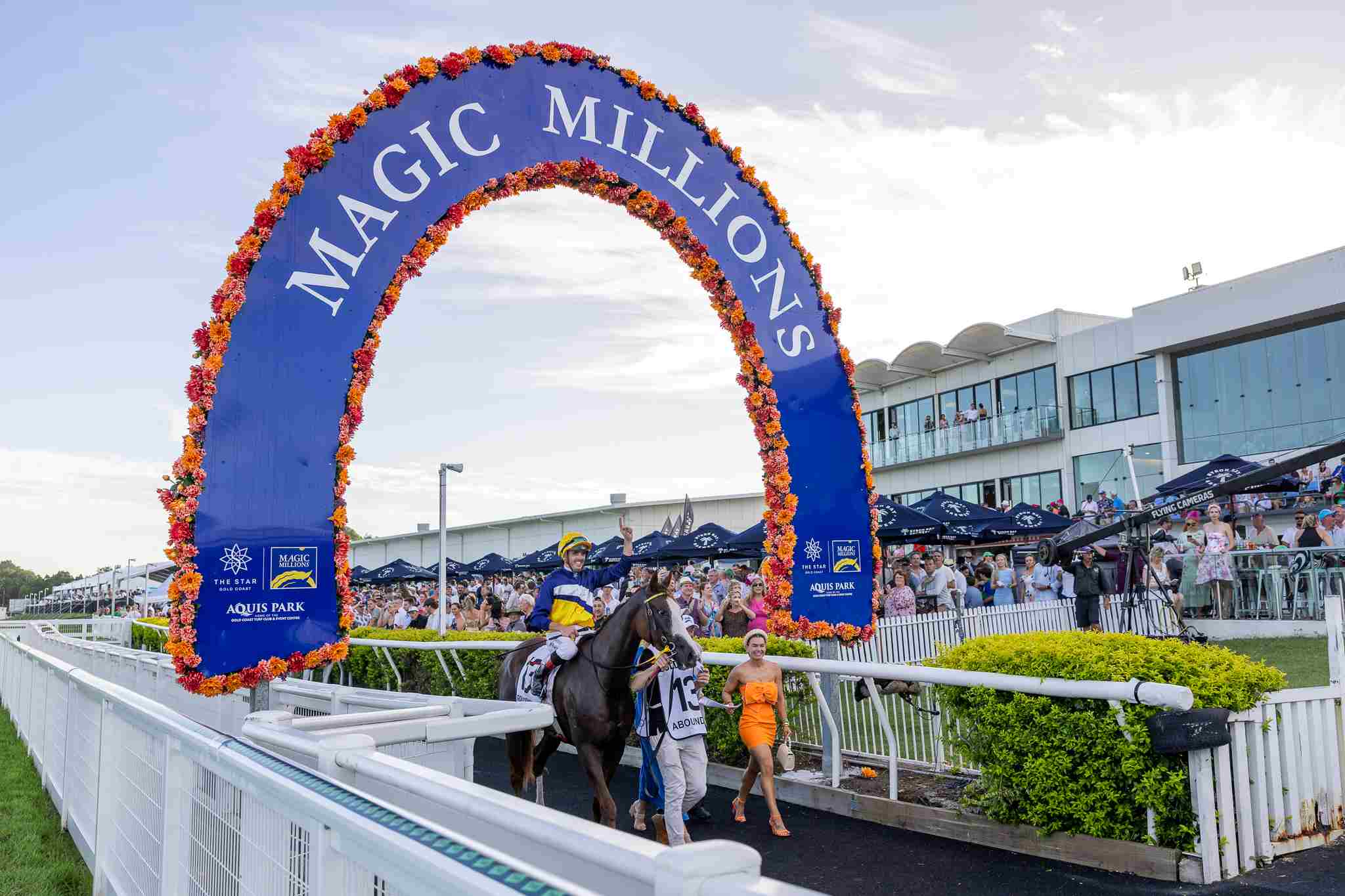 places to stay gold coast near Magic Millions raceday, with a jockey on a racehorse passing under the floral Magic Millions arch as a crowd watches